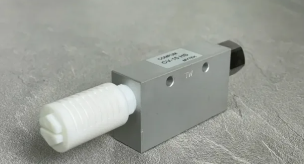 Metal rectangular actuator/valve with a white threaded plastic nozzle protruding from the left and a black connector on the right, lying on a gray surface.